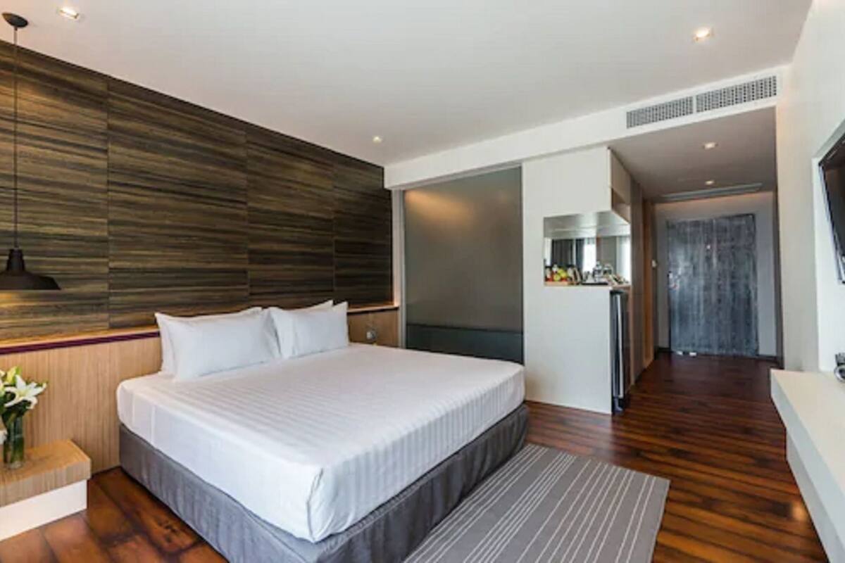 A modern hotel room with a large bed, wooden panel wall, and a small kitchenette.