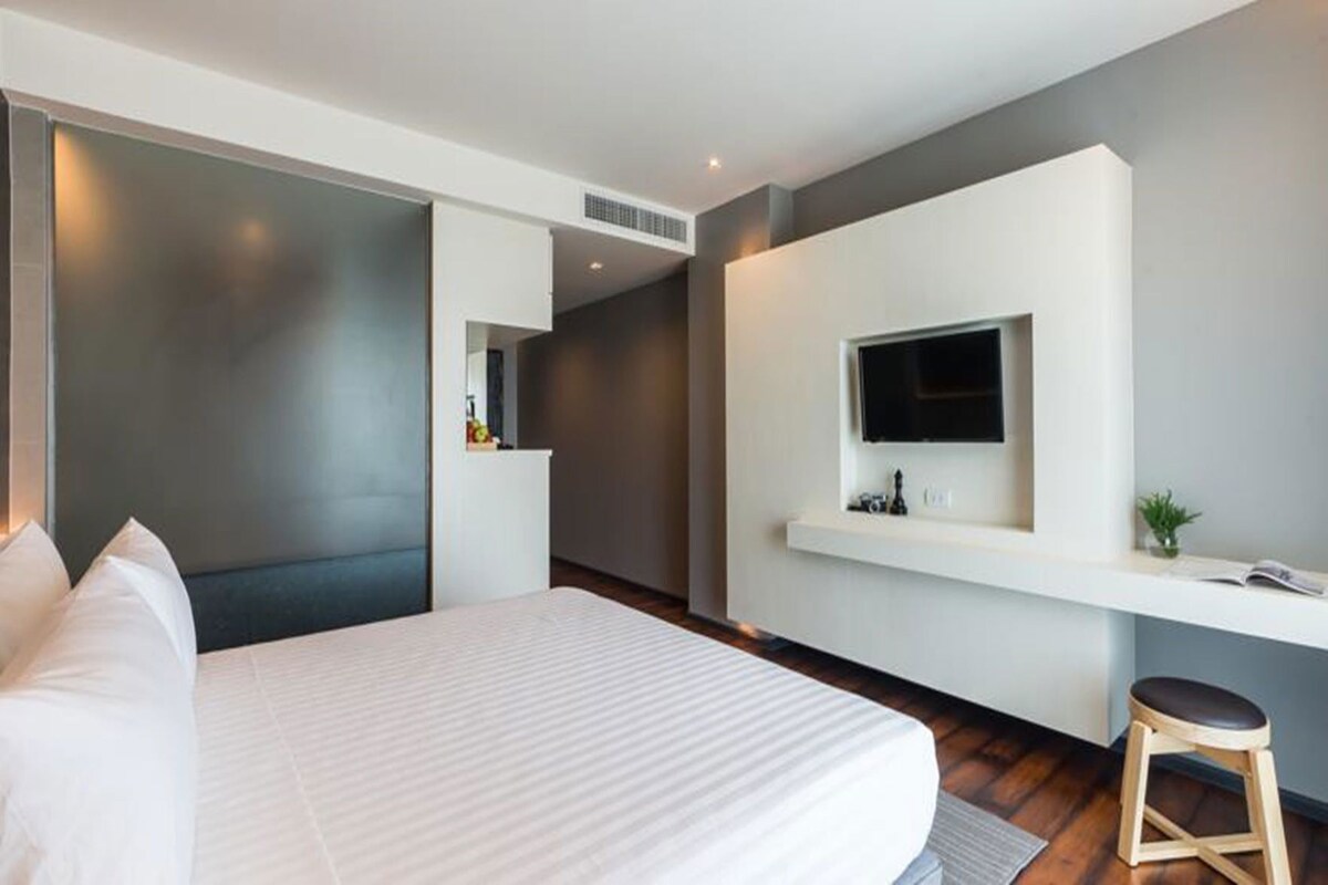 A modern hotel room with a large bed, a TV mounted on a white wall, a small stool, and a desk with a plant.