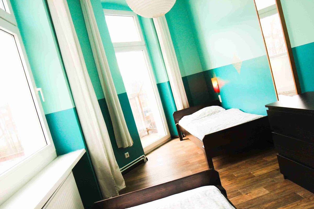 Una habitación con paredes color turquesa, suelo de madera, una cama, un aparador y una ventana con cortinas.