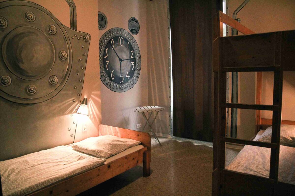 Una cama individual con estructura de madera y cabecero con remaches, un reloj de pared redondo, y una mesita con una silla.
