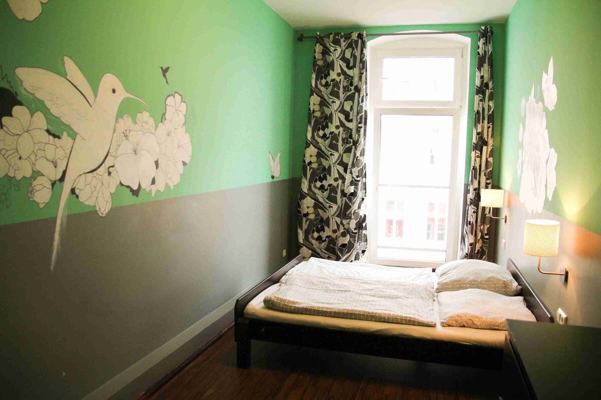 Una habitación con cama, ventana con cortinas, un cuadro floral en la pared y lámpara en la mesita de noche.