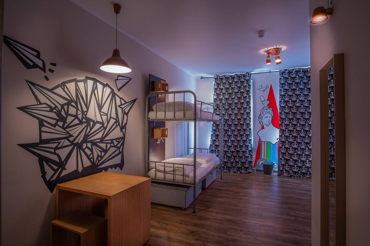 Habitación con literas, un escritorio de madera, decoración mural geométrica, cortinas estampadas y una lámpara en el techo.