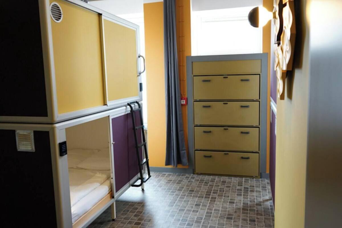 Habitación cápsula con cama doble y distribución compartida, que incluye una cama, armario y un pequeño escritorio.