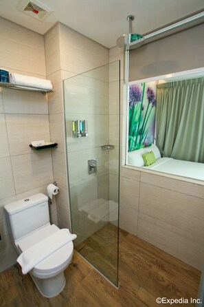 A modern bathroom with a glass shower enclosure, a toilet, and a window seat with a view of greenery. - J8 Hotel (Singapore)