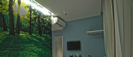 A bedroom with a bed, a desk, a TV, and a large window with a forest view.