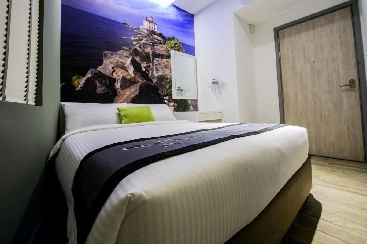 A hotel room with a bed, a painting of a lighthouse on the wall, a wooden door, and a small bedside table.
