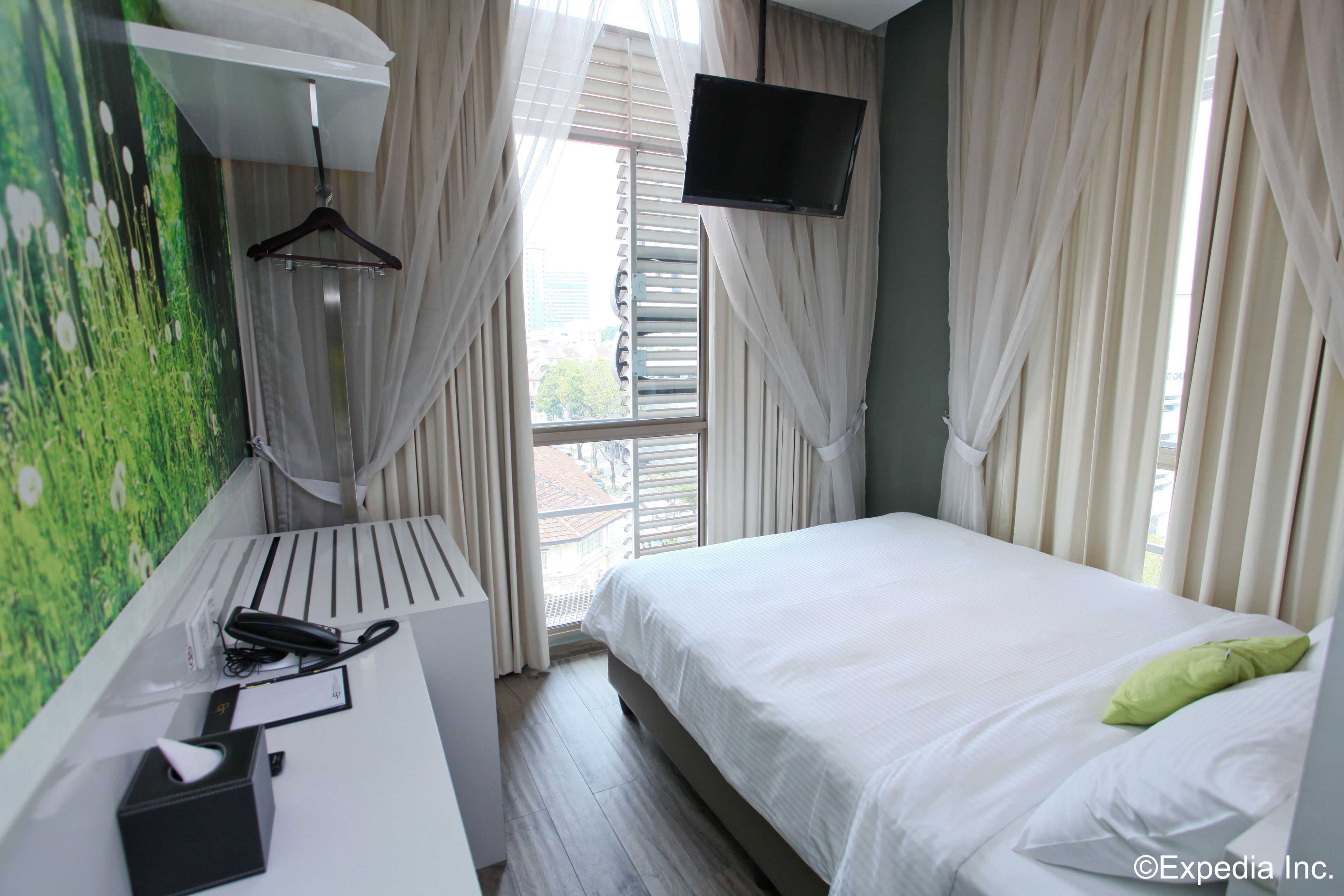 A hotel room with a bed, a desk with a telephone and a box, a television on the wall, and a window with curtains.
