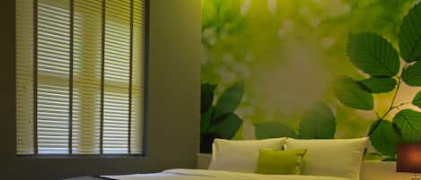 Een slaapkamer met een groot bed, een raam met zonwering, een wandlamp en een groene muur met bladmotief.