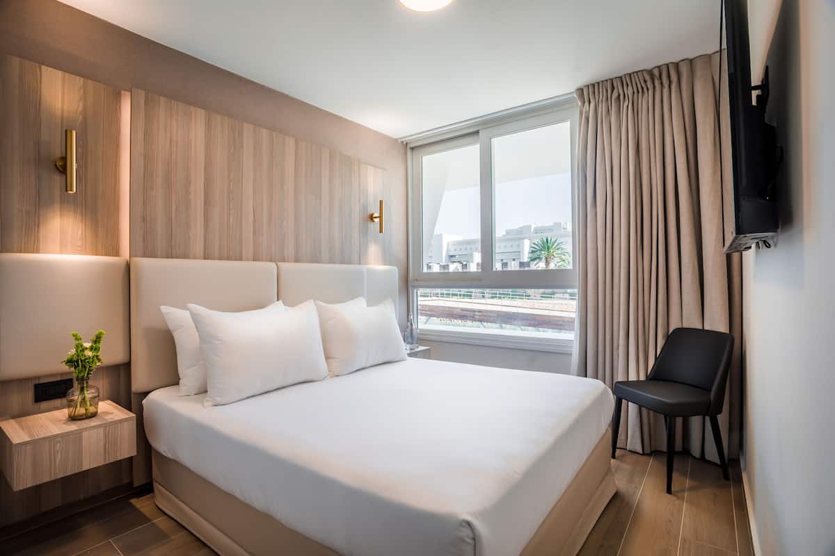 A modern hotel room with a large bed, a bedside table with a vase of flowers, a chair, and a TV mounted on the wall.