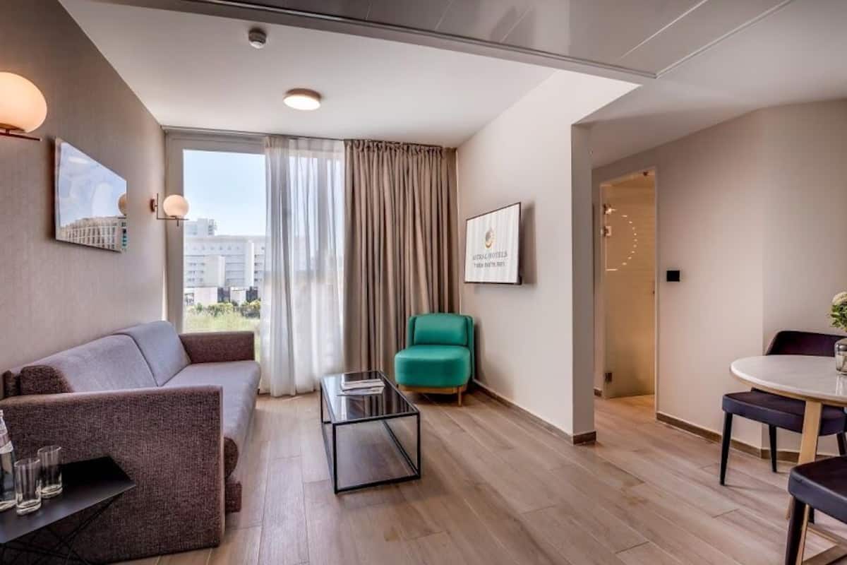 A modern hotel room with a sofa, a green armchair, a small table, and a dining area with two chairs.