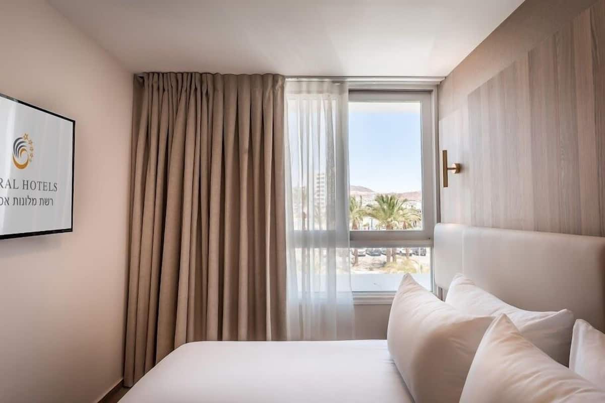 A hotel room with a bed, a window with curtains, and a view of palm trees and buildings.