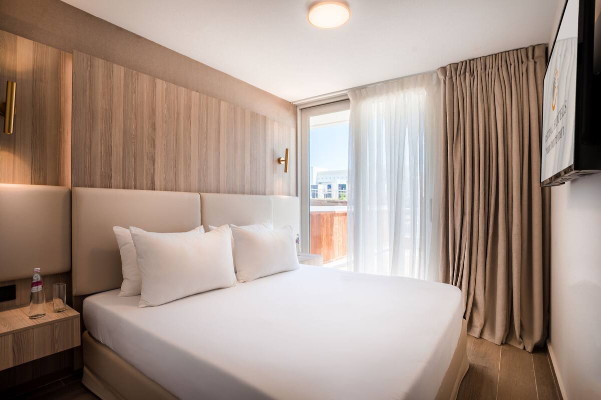 A hotel room with a large bed, a wooden headboard, a nightstand with a bottle and glass, and a TV mounted on the wall.