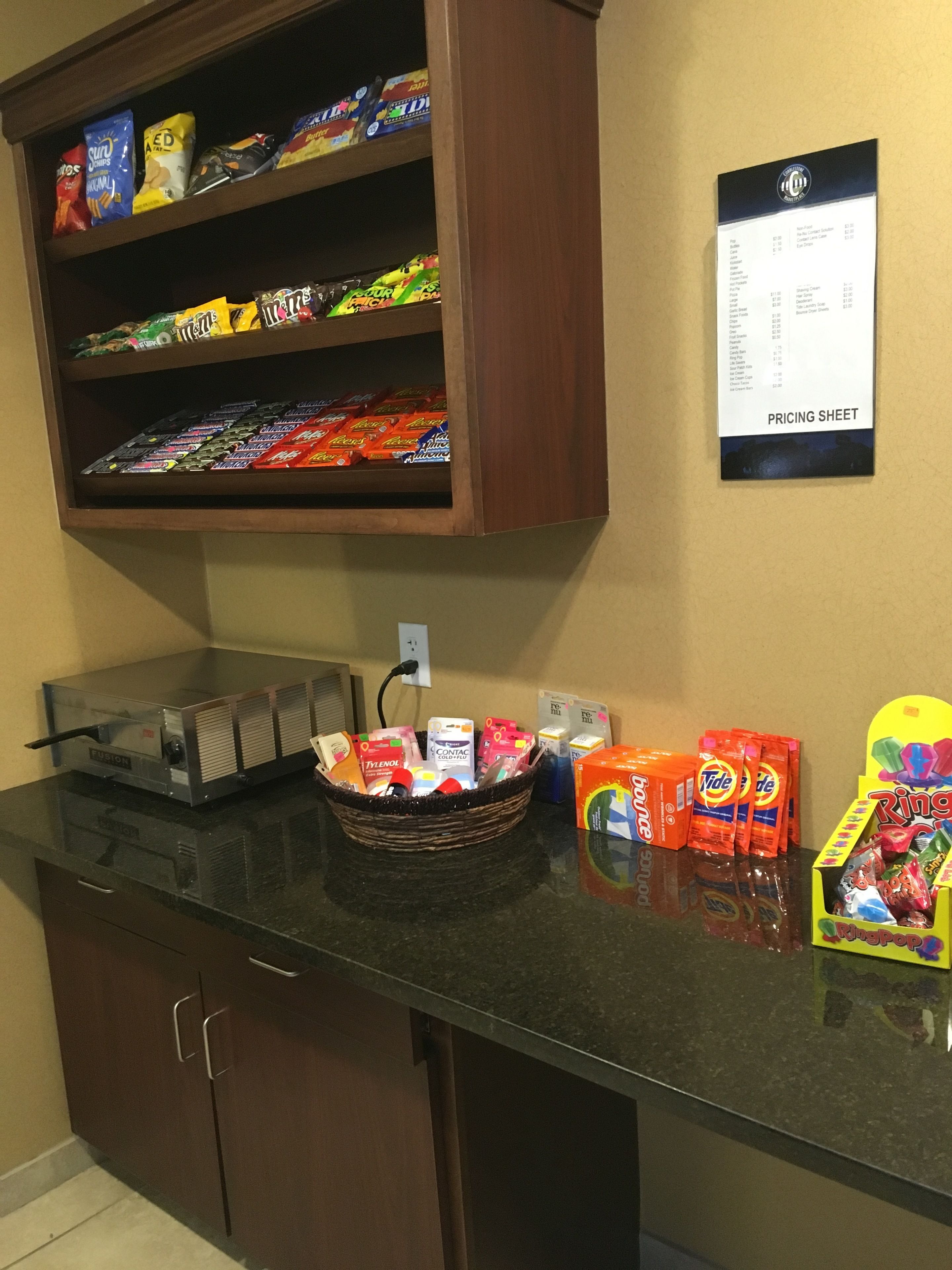A kitchenette with a refrigerator, a microwave, and a coffee maker. There is a shelf with snacks and a pricing sheet on the wall.