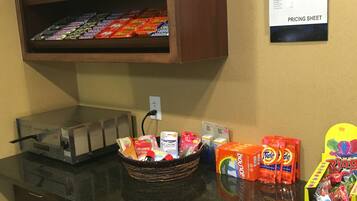 A kitchenette with a refrigerator, a microwave, and a coffee maker. There is a shelf with snacks and a pricing sheet on the wall.
