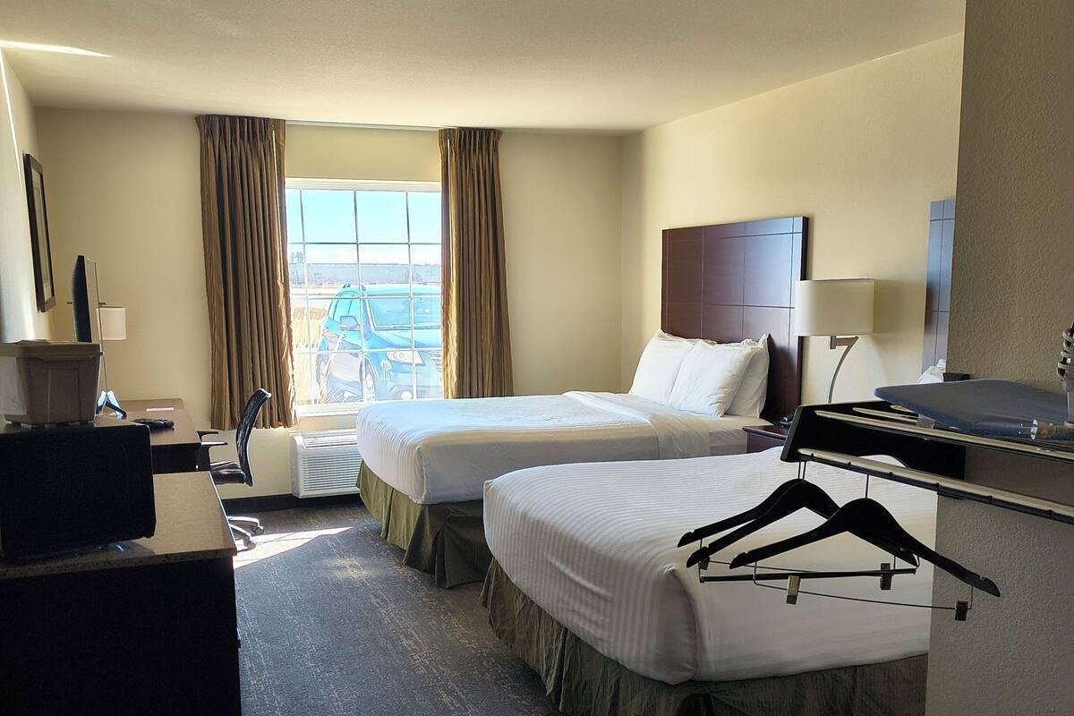 A hotel room with two beds, a desk, a chair, a TV, a lamp, and a view of the outdoors.