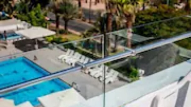 A glass-enclosed balcony with a pool, white lounge chairs, and a table with a bowl of fruit and water bottles. - Astral Nirvana Suites (Eilat)