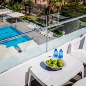 A glass-enclosed balcony with a pool, white lounge chairs, and a table with a bowl of fruit and water bottles. - Astral Nirvana Suites (Eilat)