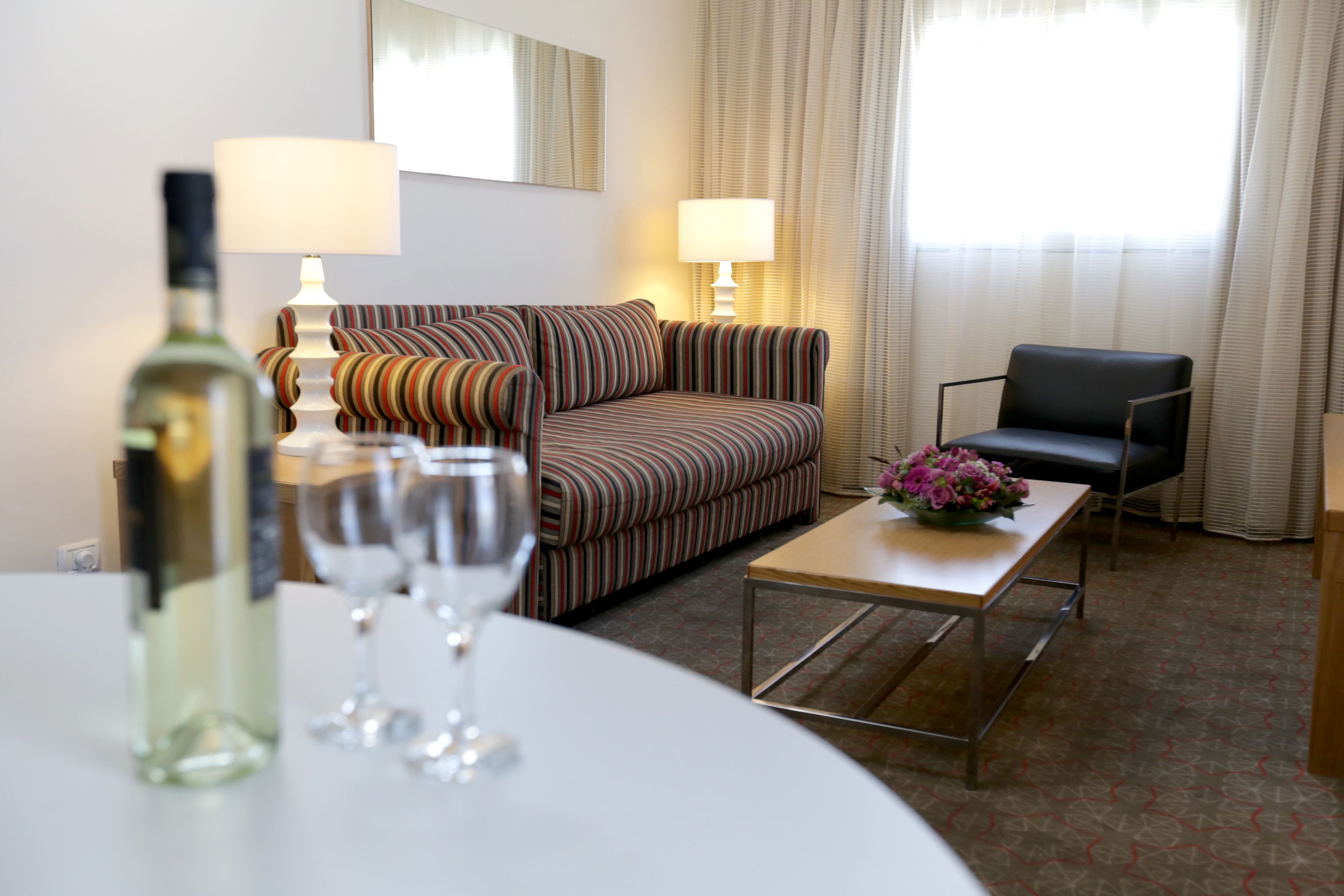 Ein Hotelzimmer mit einer gestreiften Couch, einem schwarzen Sessel, einem kleinen Tisch mit Blumenvase und einer Weinflasche.