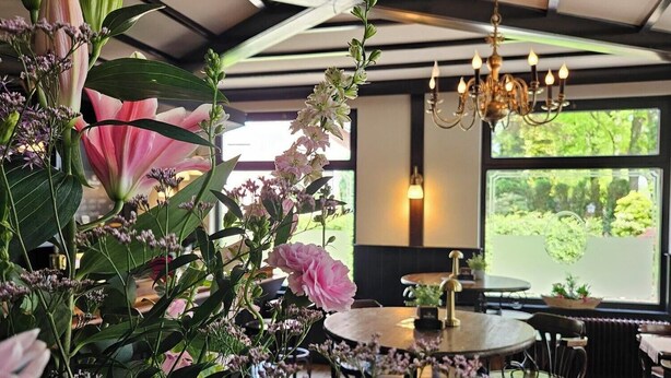 A dining area with a round table, chairs, and a chandelier. There is a large window with a view of greenery outside. - Hotel Arcense Herberg (Arcen)