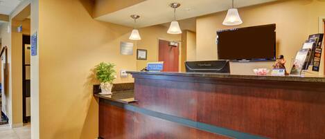 A reception desk with a wooden finish, a flat-screen TV above it, and a sign for a self-service kiosk.
