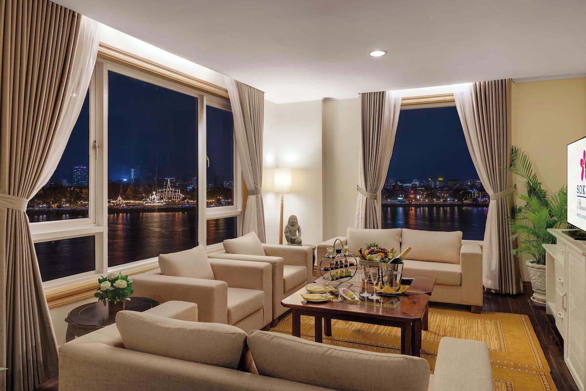 Habitación de hotel con un amplio ventanal que ofrece vistas nocturnas de la ciudad, un sofá, una mesa de centro con detalles y una pequeña estatua.