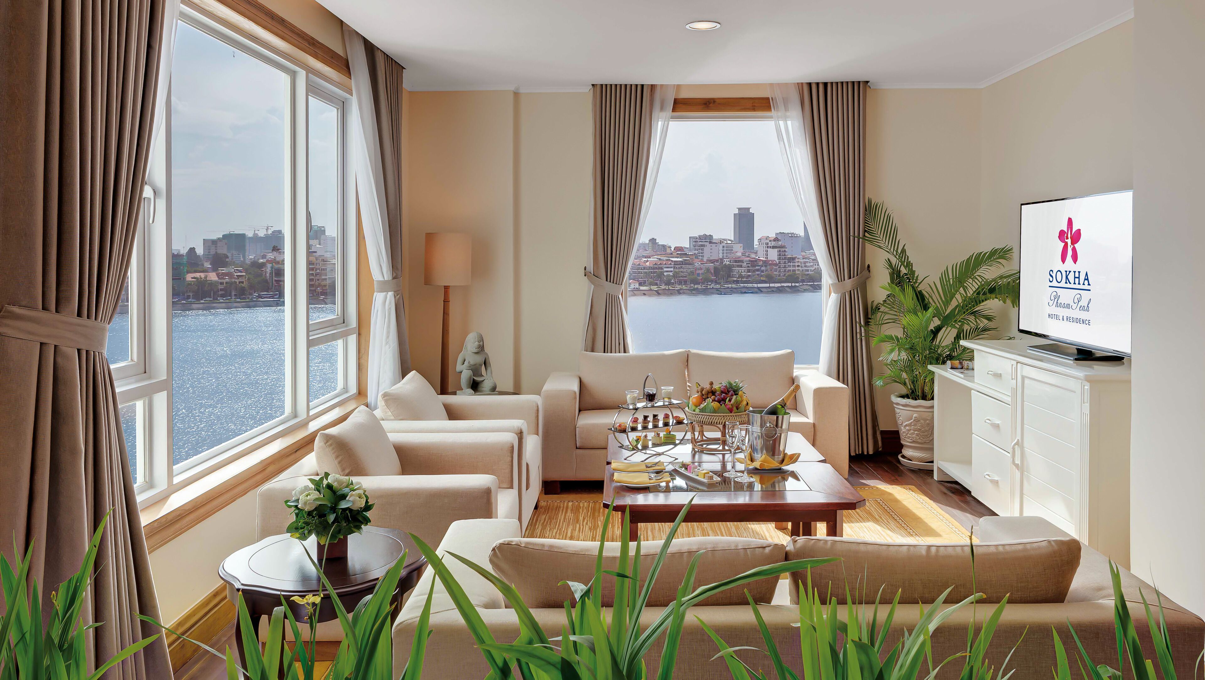 A hotel room with a city view, a sofa set, a dining table with a fruit basket, a TV, and a potted plant.
