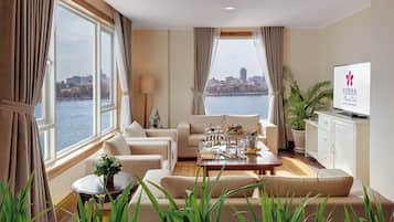 A hotel room with a city view, a sofa set, a dining table with a fruit basket, a TV, and a potted plant.
