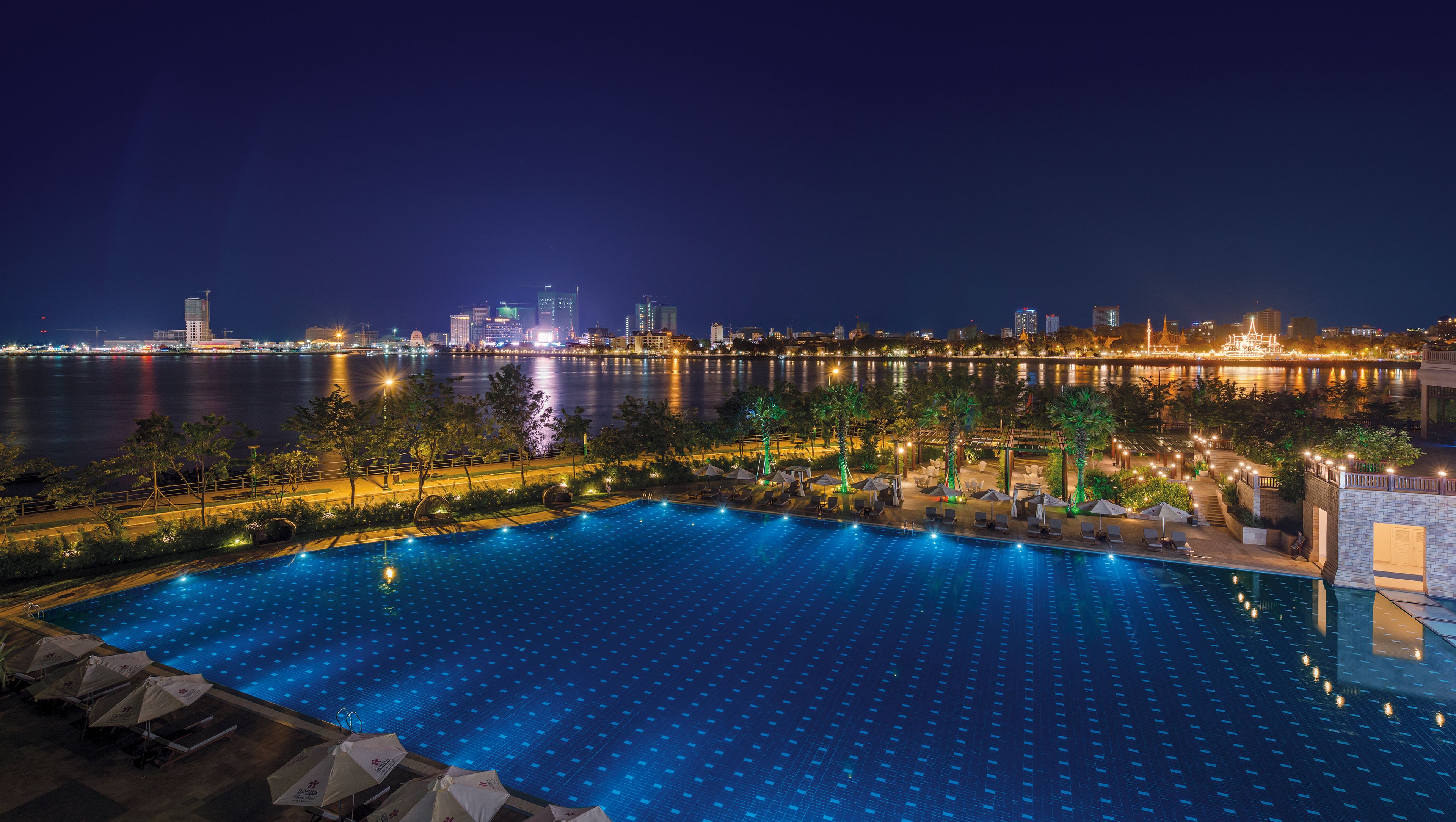 A nighttime view of a swimming pool with lounge chairs and umbrellas, overlooking a city skyline and a body of water.