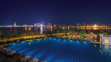A nighttime view of a swimming pool with lounge chairs and umbrellas, overlooking a city skyline and a body of water.