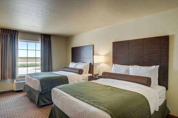 A hotel room with two beds, a large window with curtains, a wall-mounted headboard, and a bedside table with a lamp.