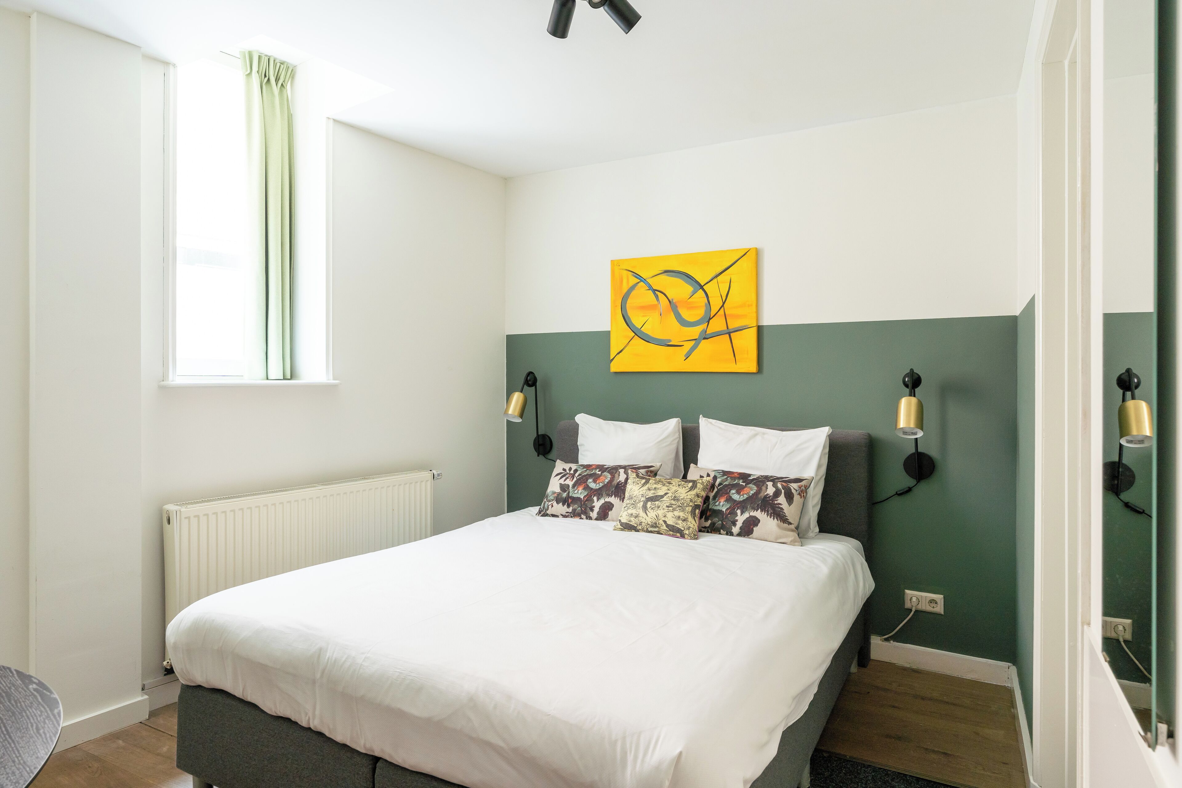 Ein ordentlich eingerichtetes Schlafzimmer mit einem Bett, einem Fenster mit Vorhängen, einem an der Wand hängenden Kunstwerk und Nachttischlampen.