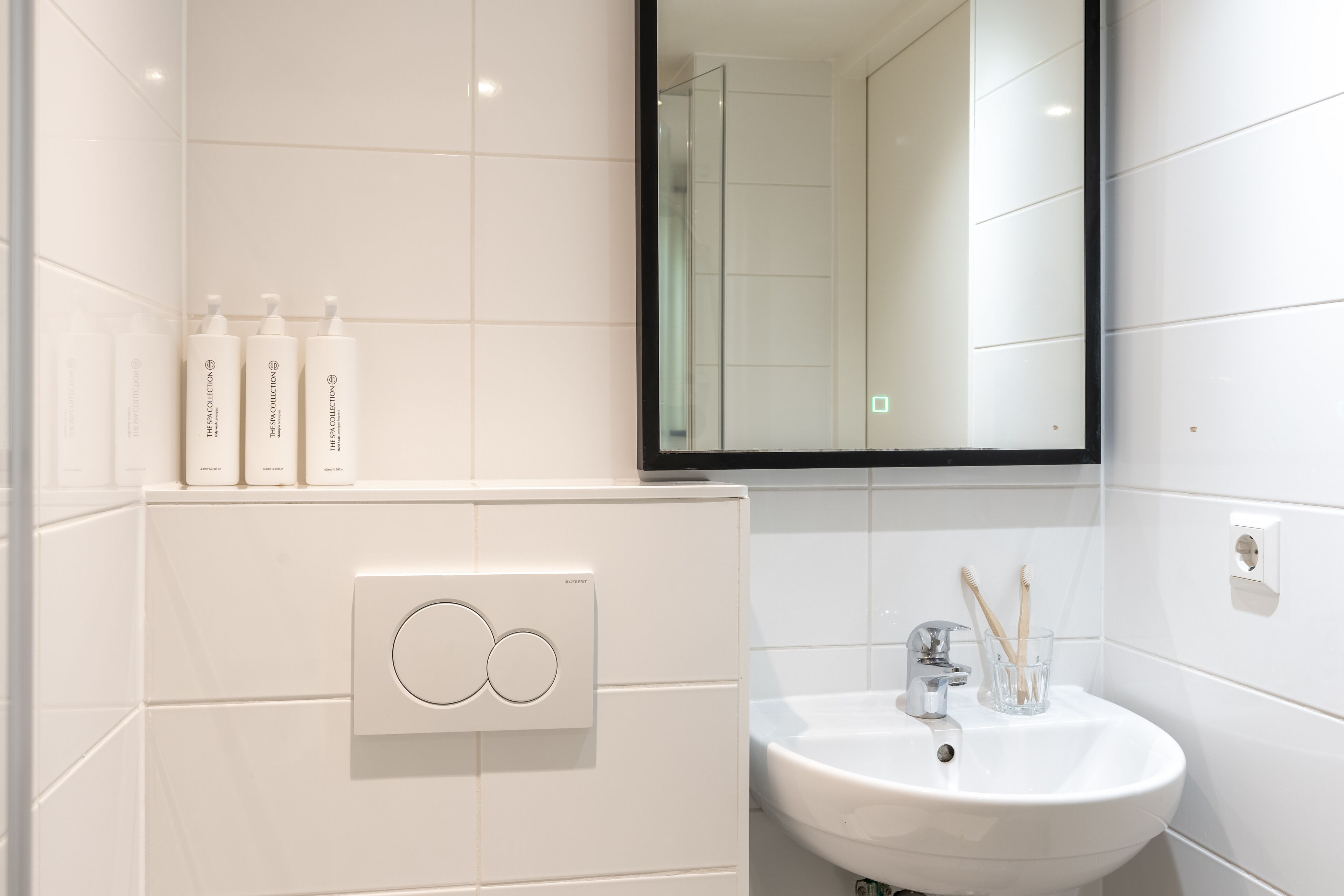 A modern bathroom with a white sink, a mirror, and three bottles of shampoo on a white cabinet.