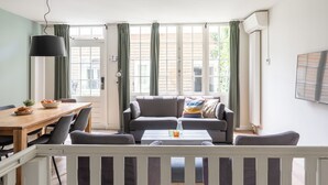 Una sala de estar con sofá, mesa de centro y mesa de comedor. Hay cortinas en las ventanas y un equipo de aire acondicionado montado en la pared.