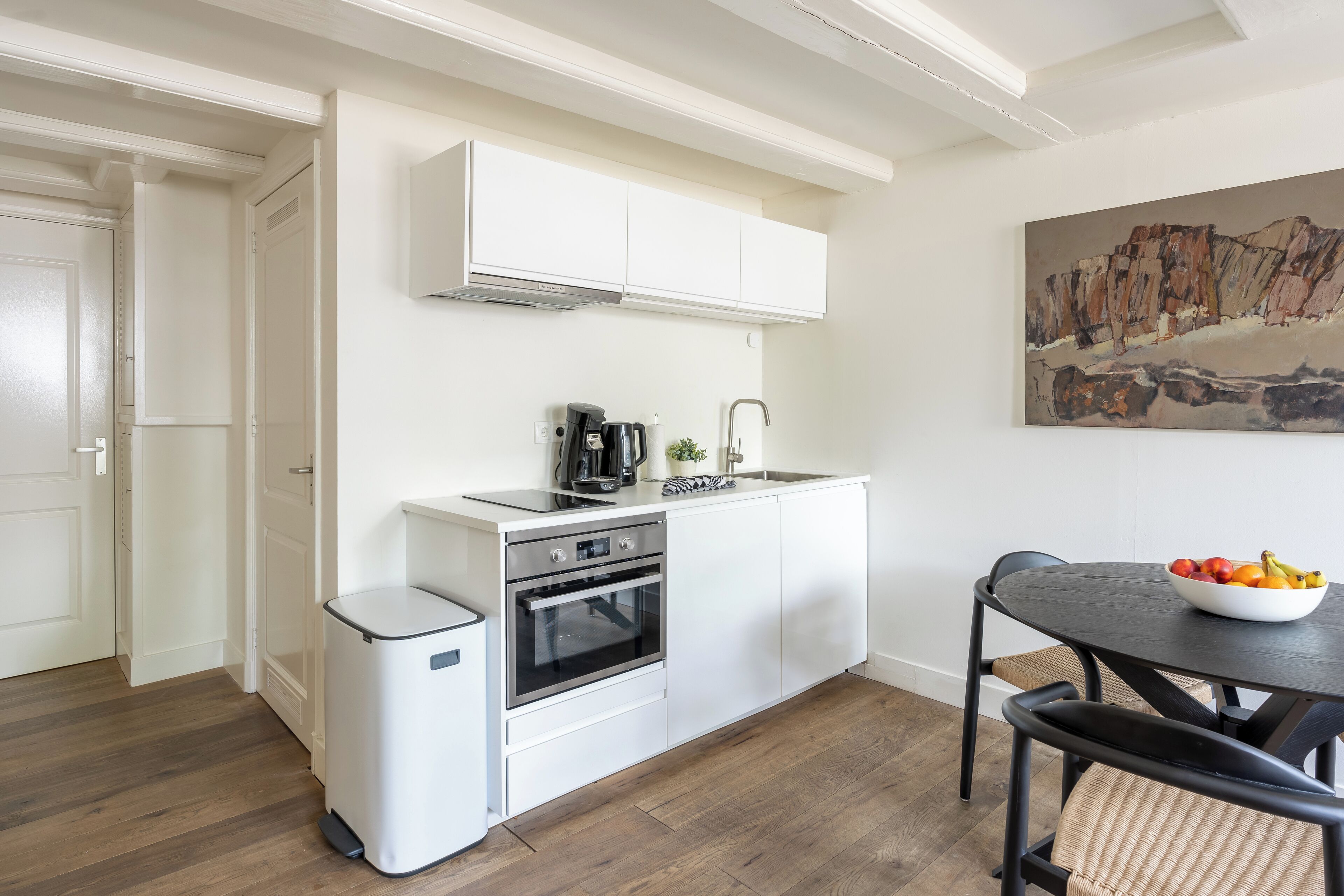 Una cucina compatta con mobili bianchi, forno integrato, macchina per caffè e una ciotola di frutta sul tavolo da pranzo.