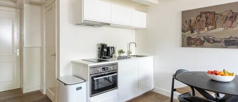 Una cucina compatta con mobili bianchi, forno integrato, macchina per caffè e una ciotola di frutta sul tavolo da pranzo.