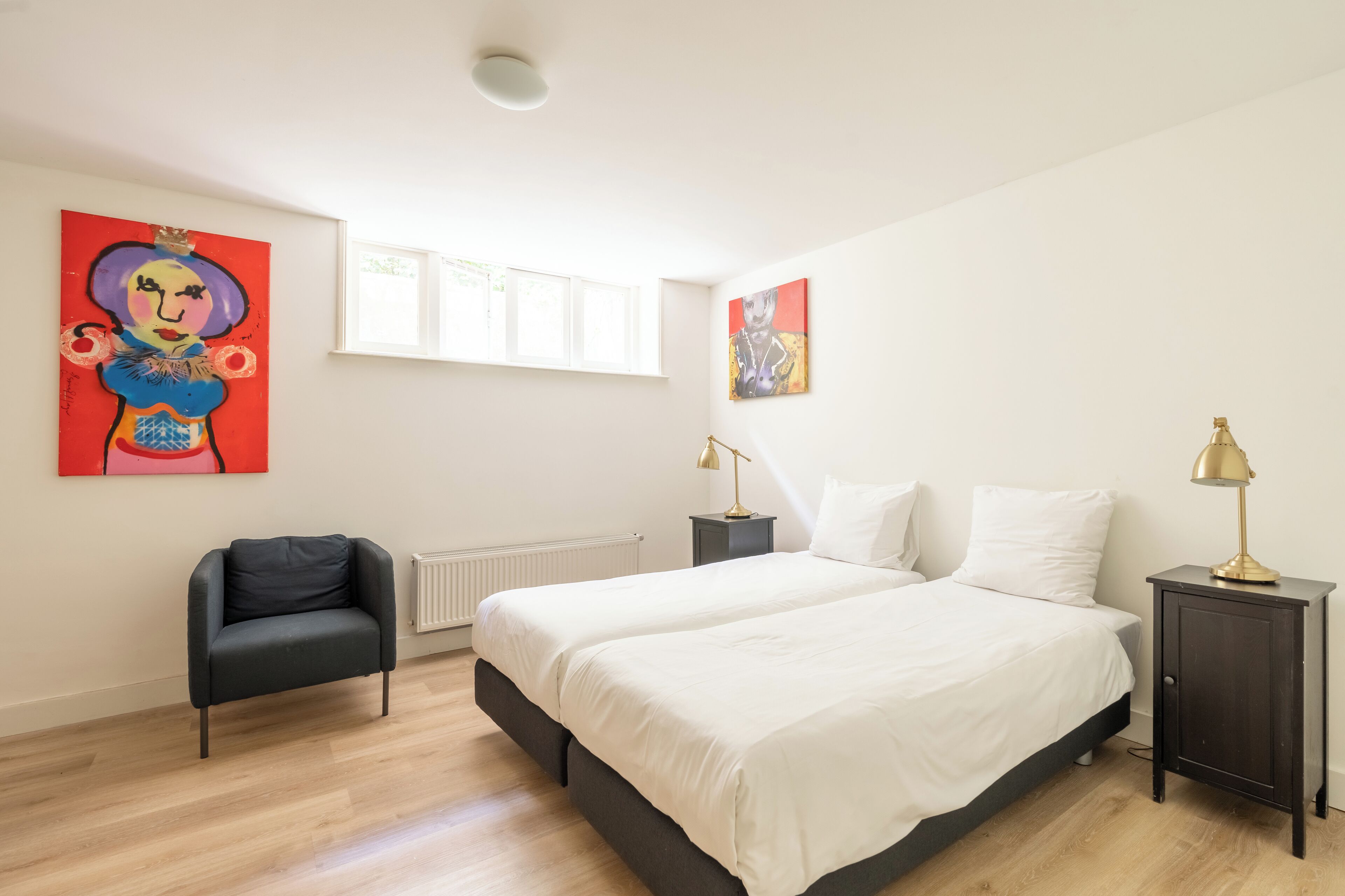 Un dormitorio con una cama doble, dos mesitas de noche, una silla y dos cuadros en la pared.