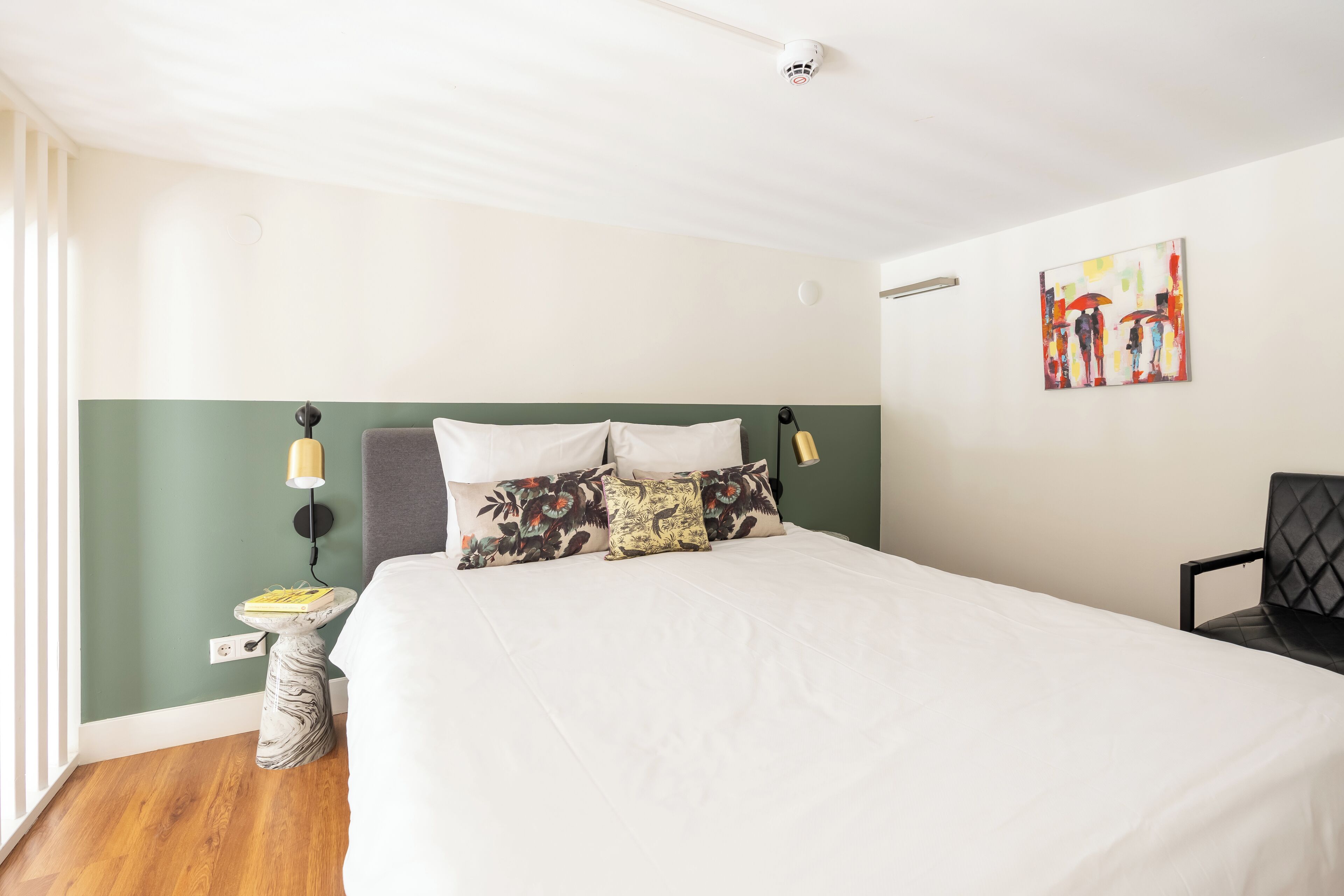 A neatly made bed with white linens, a green accent wall, and a small bedside table with a lamp.