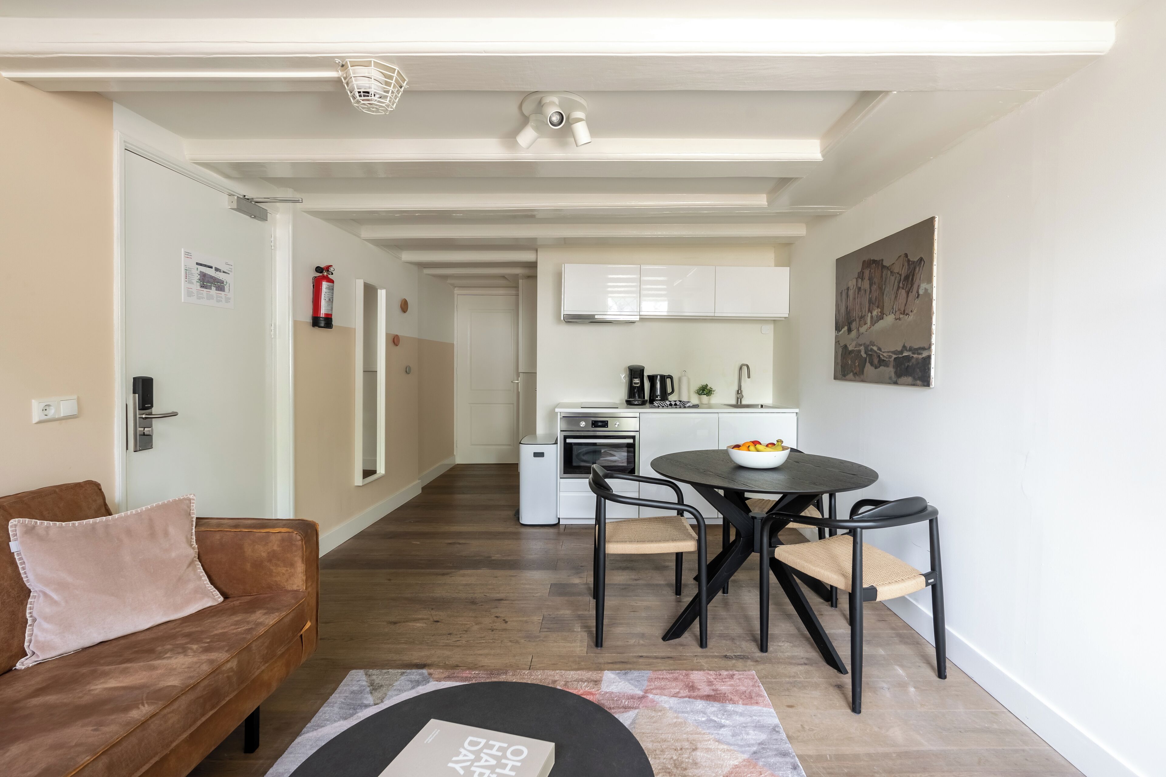 Ein modernes Wohnzimmer mit einem braunen Sofa, einem runden Esstisch mit zwei Stühlen, einer Küche im Hintergrund und einer weißen Decke mit Einbuchtungslampen.