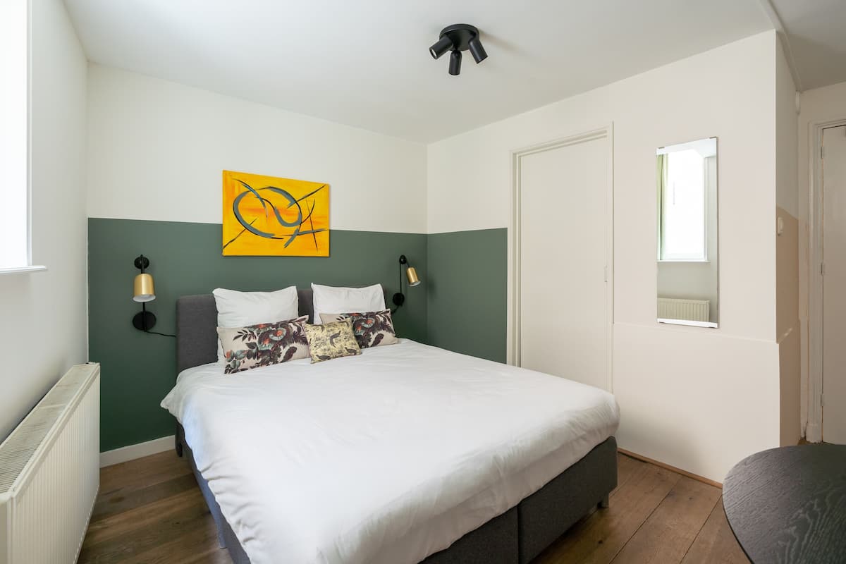 A bedroom with a bed, a wall-mounted artwork, two bedside lamps, a radiator, and a window.