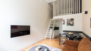 Un espacio habitable moderno con un televisor de pantalla plana montado en la pared, una escalera que lleva a un nivel superior, una zona de comedor con una mesa y sillas, y un sofá marrón.