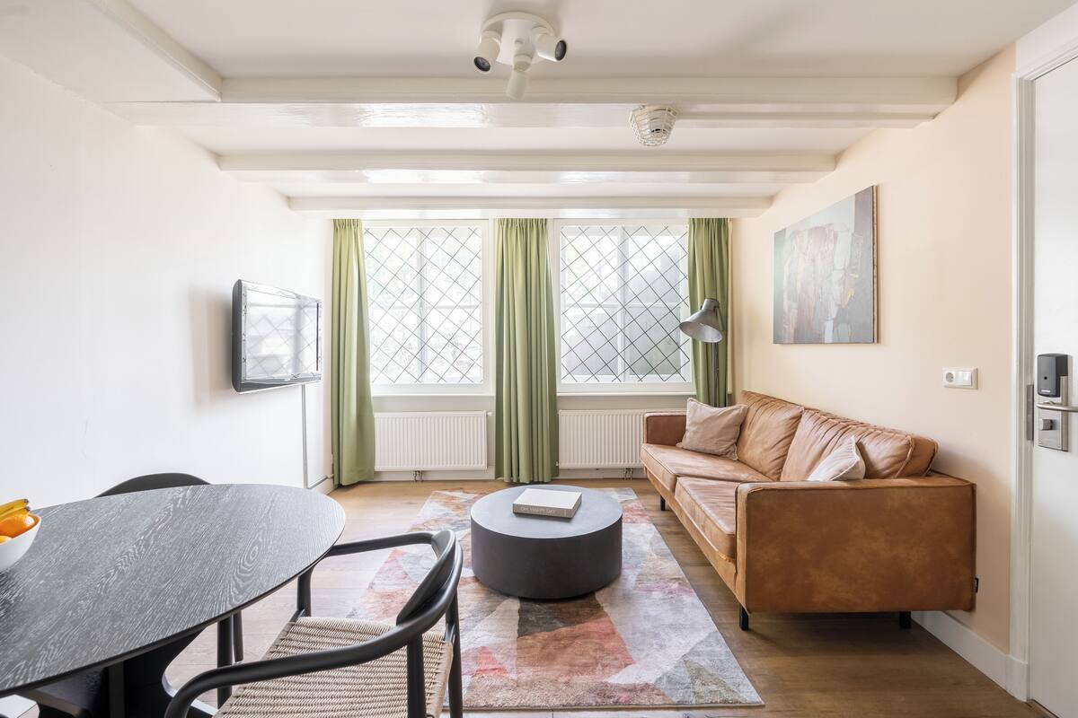 A modern living room with a round coffee table, a sofa, a dining table with chairs, and a large window with green curtains.