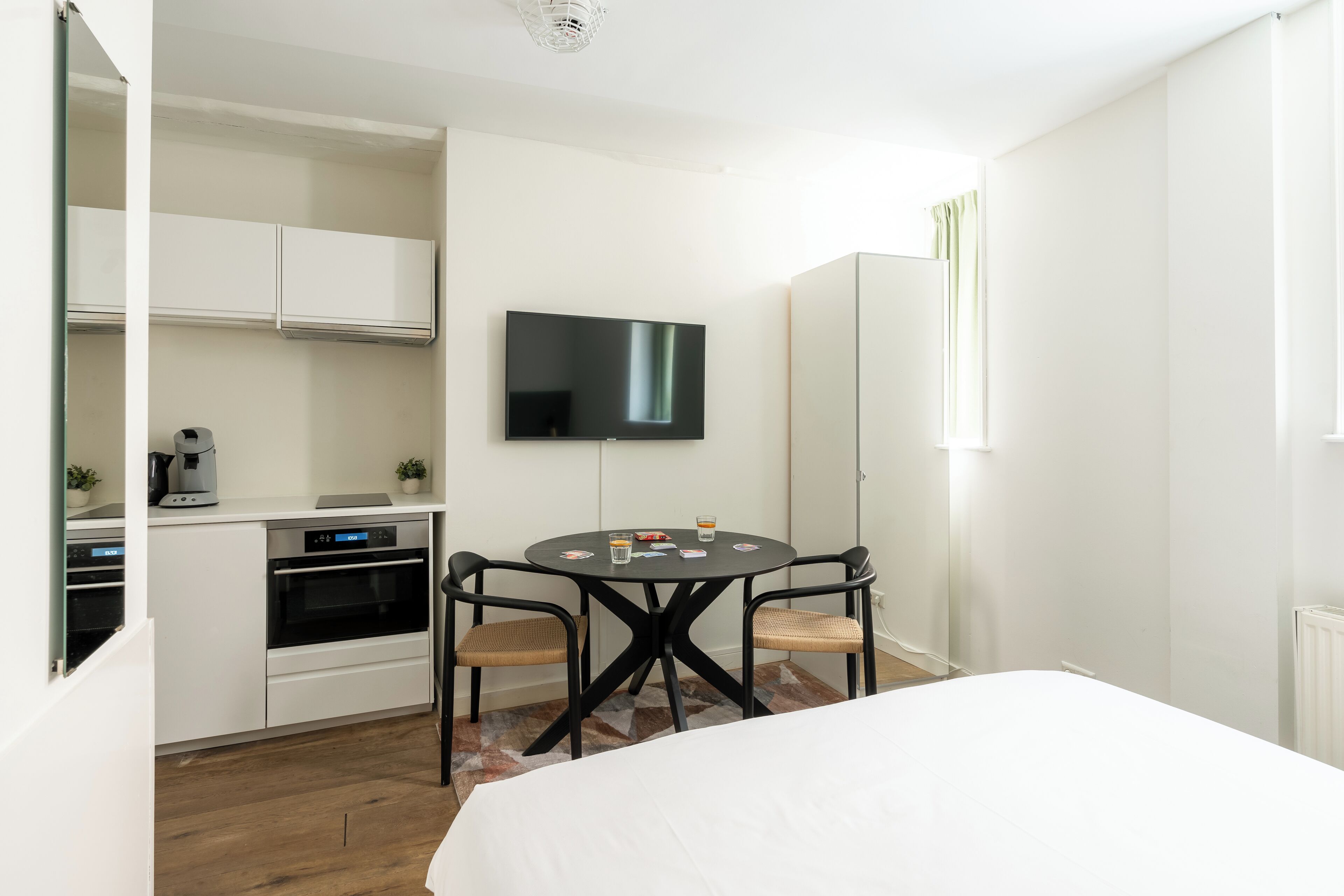 A hotel room with a bed, a dining table with chairs, a TV mounted on the wall, and a kitchenette with cabinets and appliances.