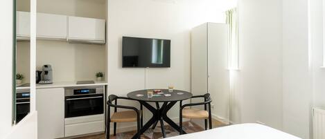 A hotel room with a bed, a dining table with chairs, a TV mounted on the wall, and a kitchenette with cabinets and appliances.