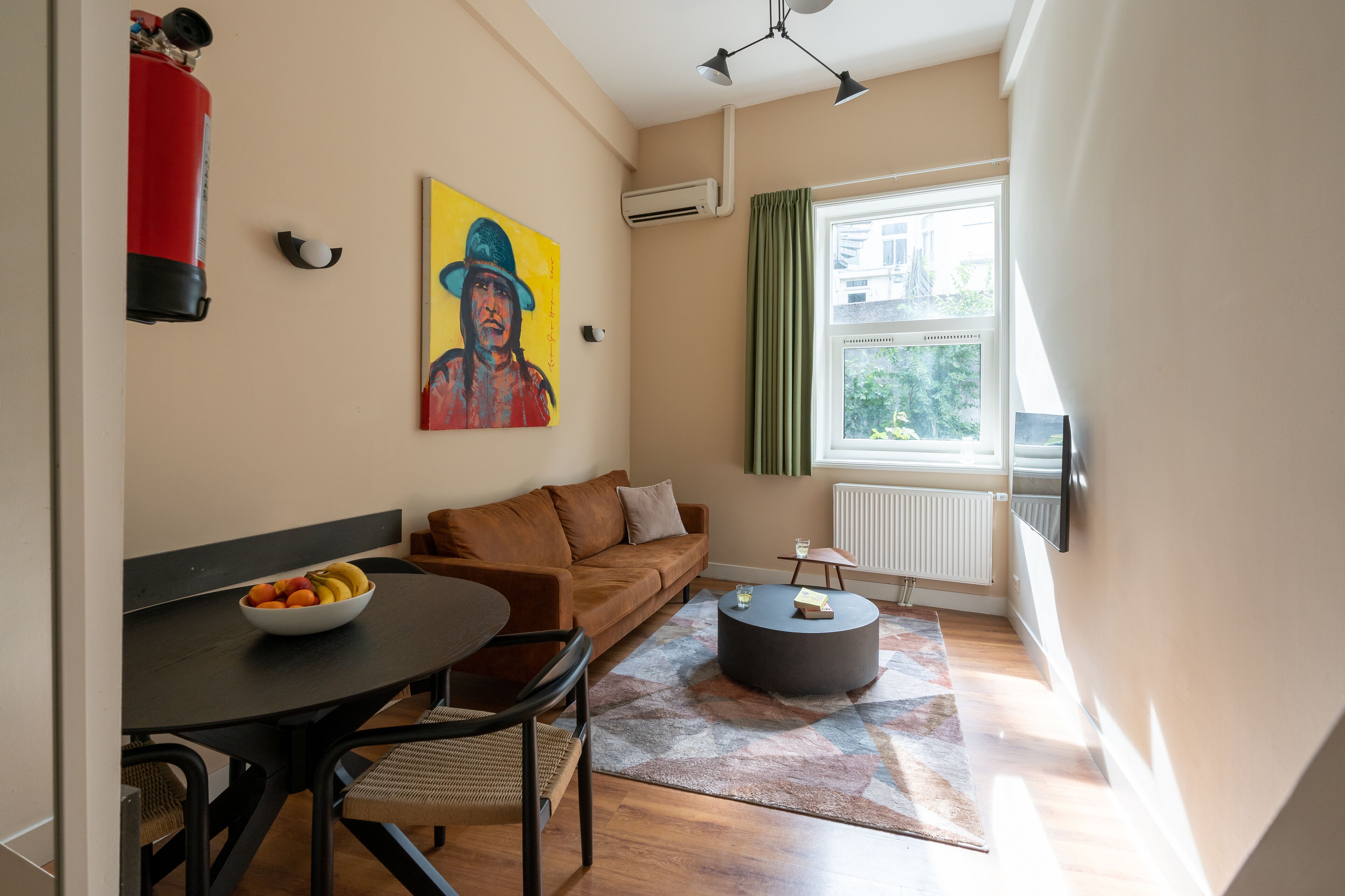 Un salon moderne avec un canapé, une table basse ronde, une table à manger sur laquelle se trouve un saladier rempli de fruits, un tableau accroché au mur et une fenêtre avec des rideaux.