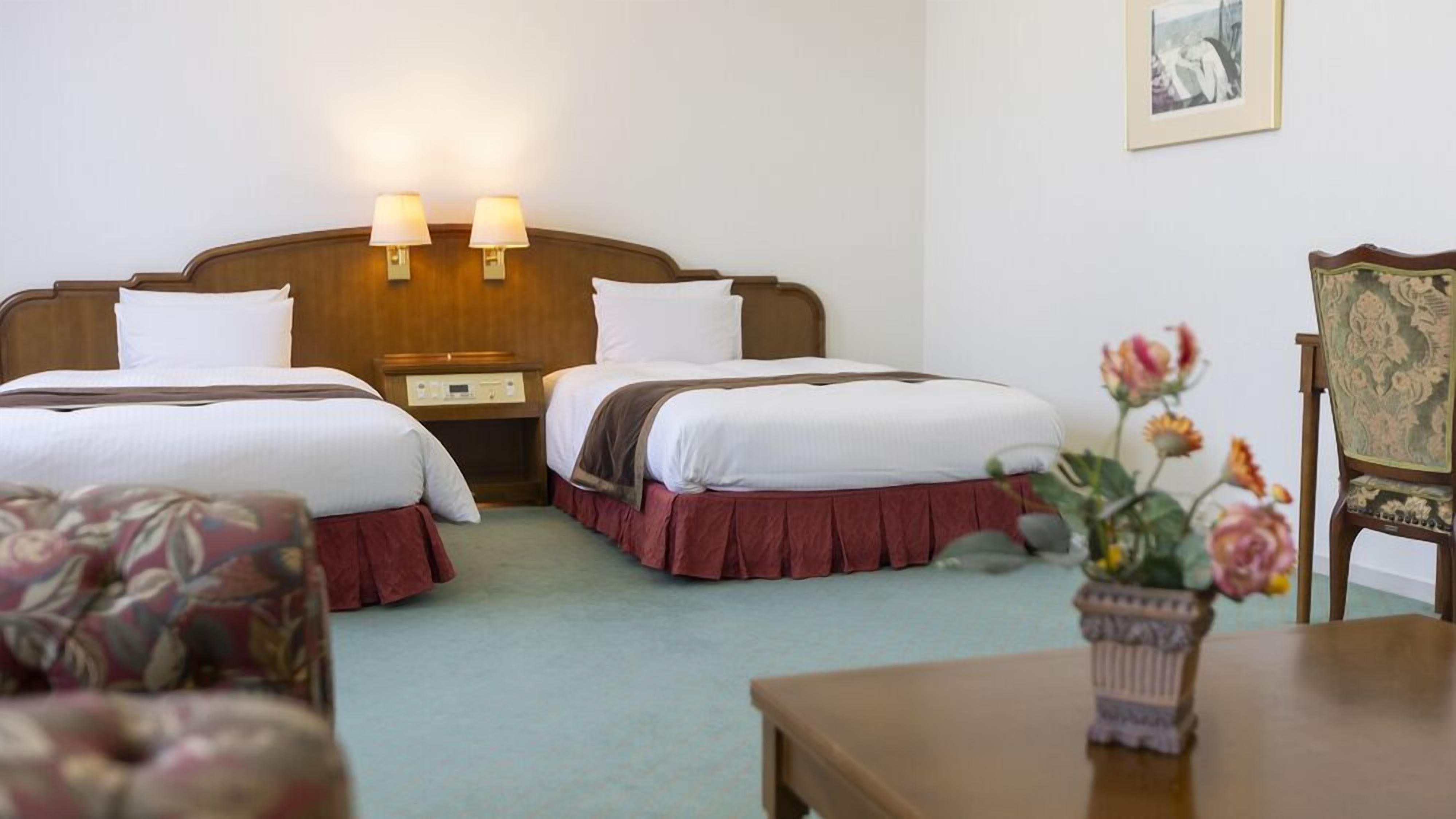 A hotel room with two beds, a nightstand, a lamp, a framed picture on the wall, and a floral arrangement on a table.