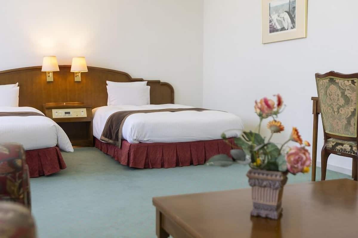 Ein Hotelzimmer mit zwei Betten, einem Nachttisch, einer Lampe, einem gerahmten Bild an der Wand und einer Blumenkomposition auf einem Tisch.