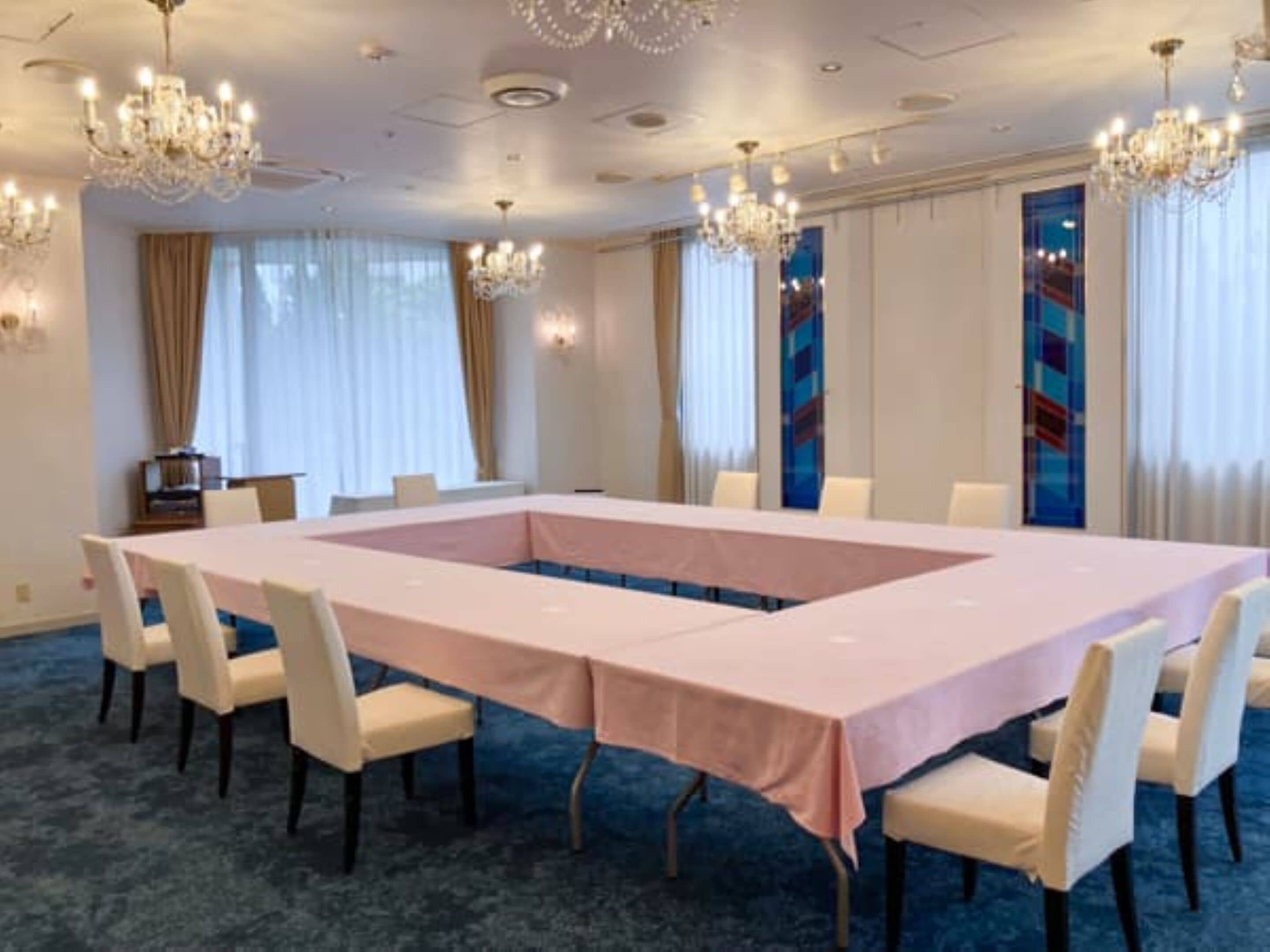 A conference room with a long table, white chairs, chandeliers, and large windows with curtains.