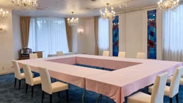 A conference room with a long table, white chairs, chandeliers, and large windows with curtains.