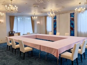 Uma sala de conferência com uma mesa comprida, cadeiras brancas, lustres e janelas grandes com cortinas.