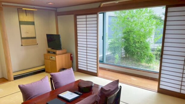 A traditional Japanese room with tatami flooring, a low table with cushions, a television on a cabinet, and a view of greenery outside.